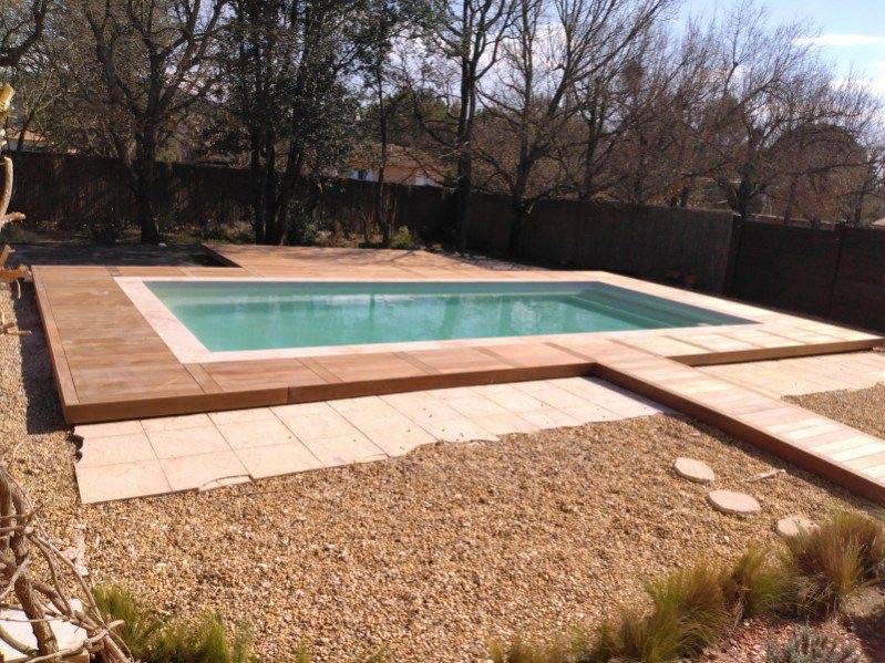 piscine coque rectangulaire installée par Generale Piscines dans le Vaucluse. Aménagement avec une grande terrasse en bois exotique et un chemin d'accès central, combinant bois et margelles claires.